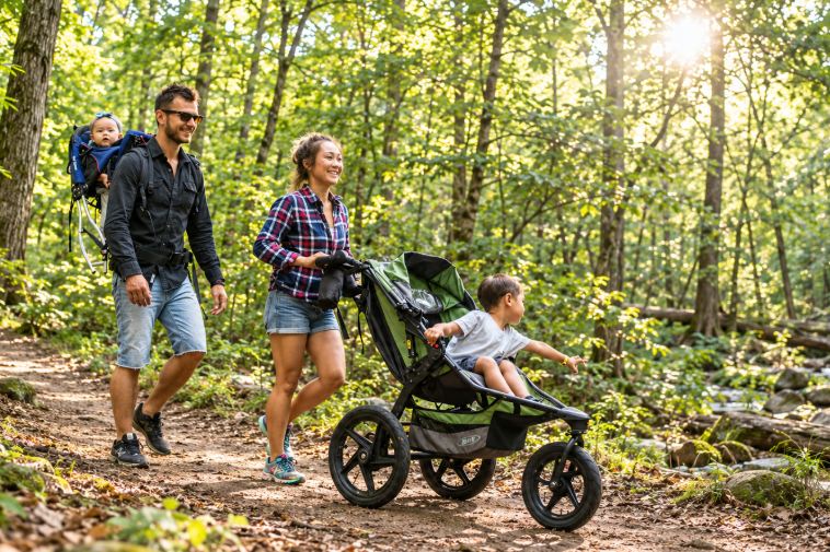A family on a forest trail with a full-size jogging stroller and a child carrier